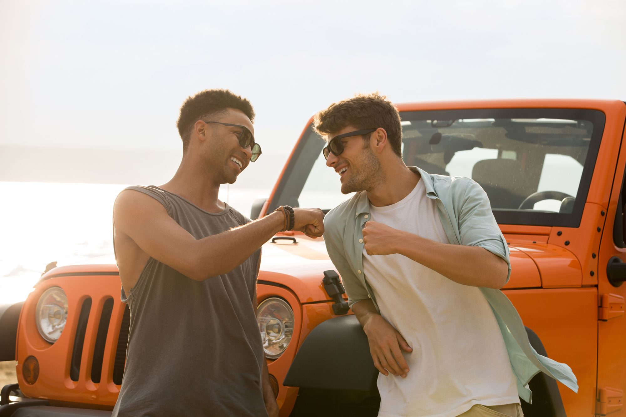 Two friends laughing and bumping fists in front of a freshly washed orange Jeep at the beach